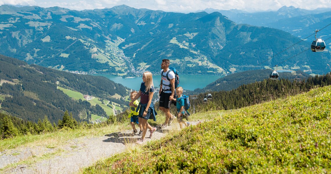 Familie wandert am Berg mit Blick auf See, Tal und Seilbahn im Hintergrund. | © Rohal, Schmittenhöhebahn AG