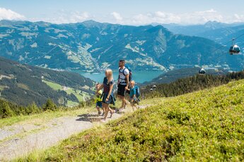 Familie wandert am Berg mit Blick auf See, Tal und Seilbahn im Hintergrund. | © Rohal, Schmittenhöhebahn AG