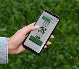 Hand holding a smartphone displaying the digital Nationalpark SommerCard, QR code visible, green meadow. | © Ferienregion Nationalpark Hohe Tauern