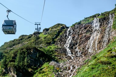 Seilbahn fährt entlang eines grünen Berghangs mit rauschendem Wasserfall in der Weißsee Gletscherwelt. | © Ferienregion Nationalpark Hohe Tauern