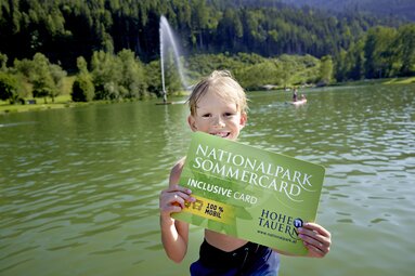 Kind mit der Nationalpark SommerCard vor einem idyllischen See mit Wald im Hintergrund. | © Ferienregion Nationalpark Hohe Tauern - Michael Huber