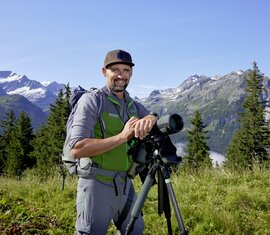 A smiling national park ranger with binoculars amidst an alpine landscape in the sunshine. | © Ferienregion Nationalpark Hohe Tauern GmbH
