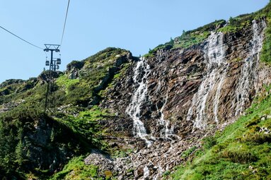 Seilbahn fährt entlang eines grünen Berghangs mit rauschendem Wasserfall in der Weißsee Gletscherwelt. | © Ferienregion Nationalpark Hohe Tauern