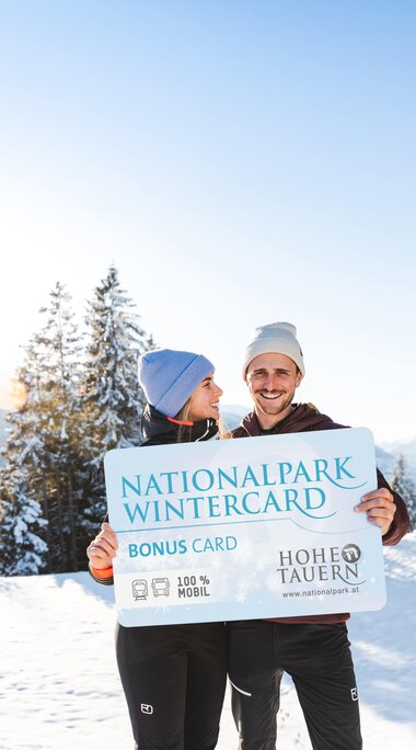 Ein lächelndes Paar hält eine große Nationalpark WinterCard vor einer winterlichen Berglandschaft. | © Ferienregion Nationalpark Hohe Tauern - Mathäus Gartner