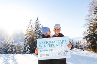 Ein lächelndes Paar hält eine große Nationalpark WinterCard vor einer winterlichen Berglandschaft. | © Ferienregion Nationalpark Hohe Tauern - Mathäus Gartner