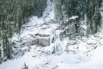 Verschneite Krimmler Wasserfälle umgeben von dichten Tannenwäldern mit einer Person im Vordergrund. | © Daniel Breuer