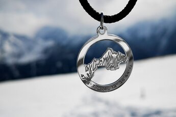 Anhänger mit Bergsilhouette und Koordinaten vor einer verschwommenen, schneebedeckten Berglandschaft. | © Gipfelband