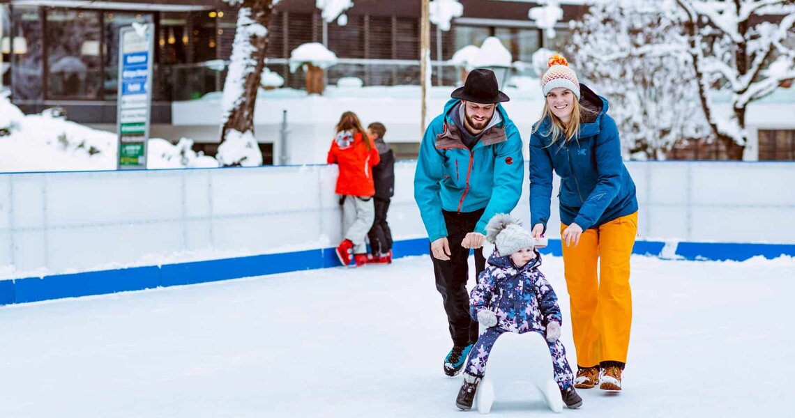 Familie schiebt ein Kleinkind mit einer Eislaufhilfe auf einer winterlichen Eislaufbahn.