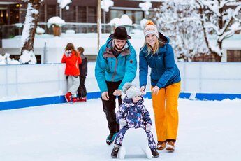 Familie schiebt ein Kleinkind mit einer Eislaufhilfe auf einer winterlichen Eislaufbahn.