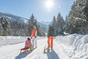 Familie mit Schlitten auf verschneitem Weg, umgeben von Bergen und Sonnenschein. | © Tourismusbüro Rauris - Fotostudio Creatina