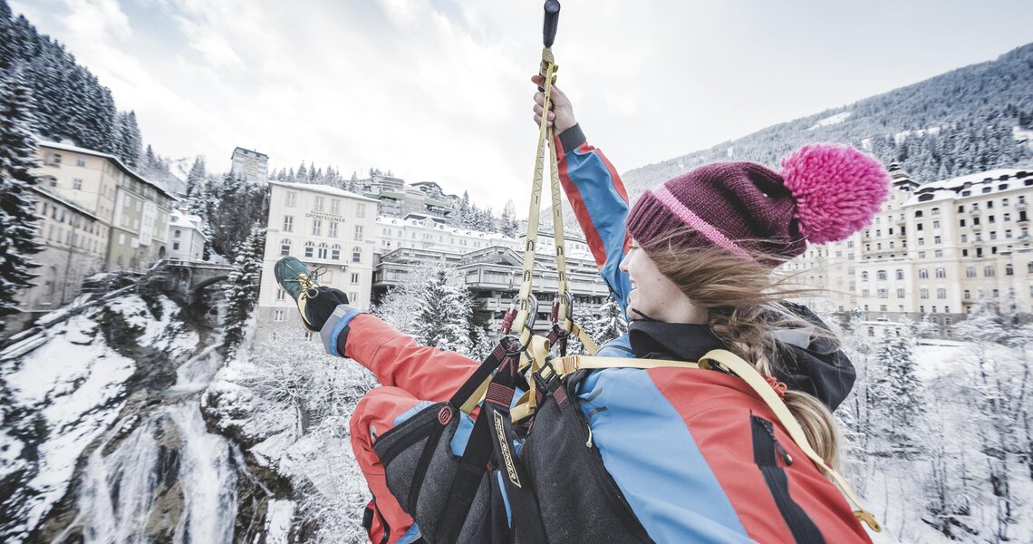 Person fährt mit einer Zipline über eine verschneite Landschaft mit Gebäuden und einem Wasserfall. | © Gasteinertal Tourismus GmbH - Klaus Listl
