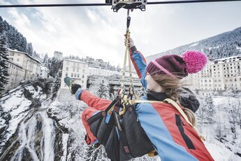 Person fährt mit einer Zipline über eine verschneite Landschaft mit Gebäuden und einem Wasserfall. | © Gasteinertal Tourismus GmbH - Klaus Listl