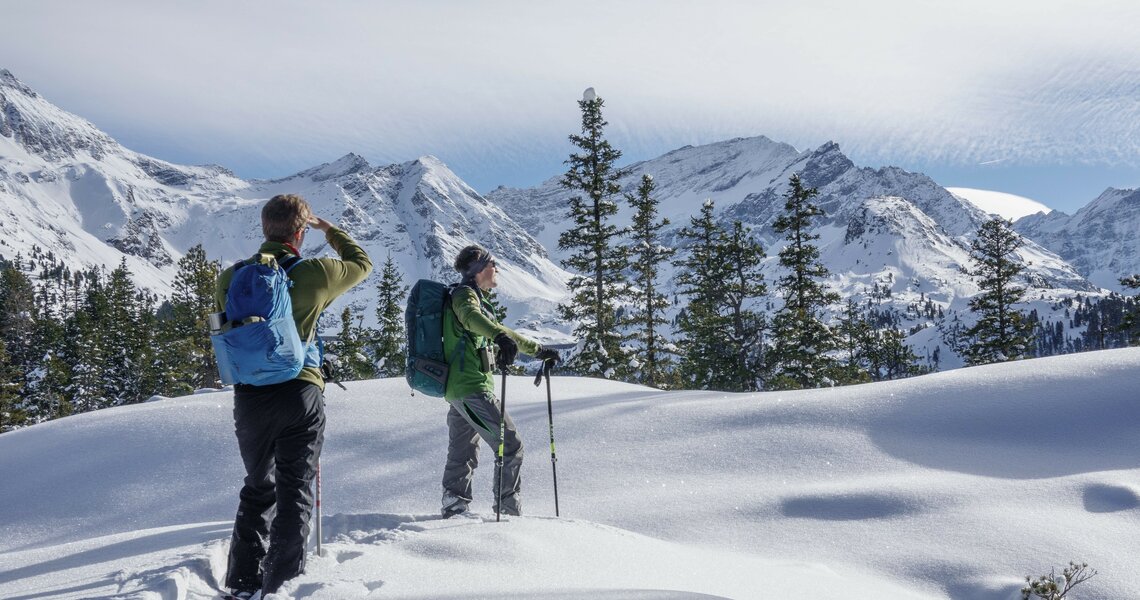 Two snowshoe hikers enjoying a snowy mountain landscape with fir trees and a sunny sky. | © Helga Baldinger