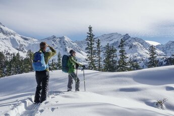 Zwei Schneeschuhwanderer genießen eine verschneite Berglandschaft mit Tannen und sonnigem Himmel. | © Helga Baldinger