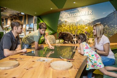 Familie erkundet interaktive Ausstellung zum Almleben mit Tischdisplay und Tierpräparaten. | © zangl.ultd-Franz Neumayr-Leo