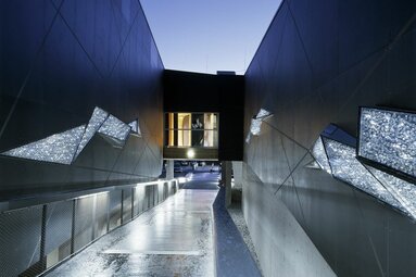 Moderner Zugang mit beleuchteten Wänden, Glasfenstern und einer Brücke bei Dämmerung. | ©  Paul Ott