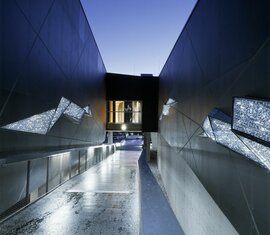 Moderner Zugang mit beleuchteten Wänden, Glasfenstern und einer Brücke bei Dämmerung. | ©  Paul Ott