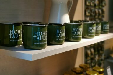 Grüne Tassen mit der Aufschrift "Hohe Tauern" stehen auf einem weißen Regal. | © Nationalparkzentrum Hohe Tauern GmbH