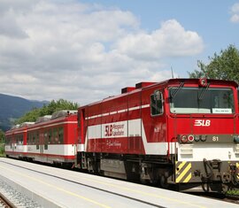 Rote Pinzgauer Lokalbahn steht an einem Bahnhof vor einer Berglandschaft bei bewölktem Himmel | © Wikipedia - Herbert Ortner