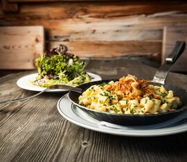 Kässpätzle in einer Pfanne mit Röstzwiebeln, serviert mit frischem Salat auf einem Holztisch | © SalzburgLand Tourismus 