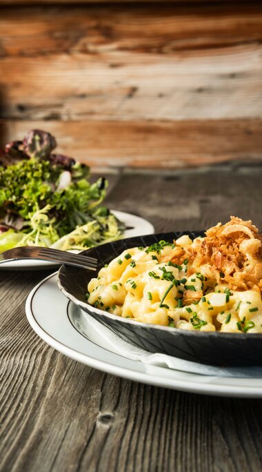 Kässpätzle in einer Pfanne mit Röstzwiebeln, serviert mit frischem Salat auf einem Holztisch | © SalzburgLand Tourismus 