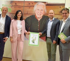 Vier Personen präsentieren ein Plakat vor einem Porträt von Marielouise Jordan in einer Ausstellung.