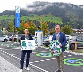 Zwei Männer präsentieren Schilder vor Ladestationen für Elektroautos mit Berglandschaft im Hintergrund.