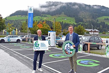 Zwei Männer präsentieren Schilder vor Ladestationen für Elektroautos mit Berglandschaft im Hintergrund.