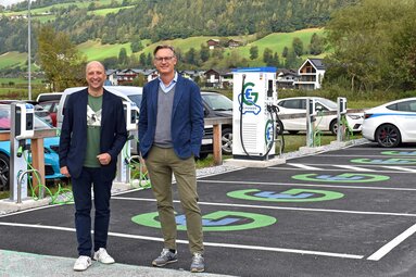 Zwei Männer stehen vor Ladestationen für Elektroautos auf einem Parkplatz in ländlicher Umgebung.