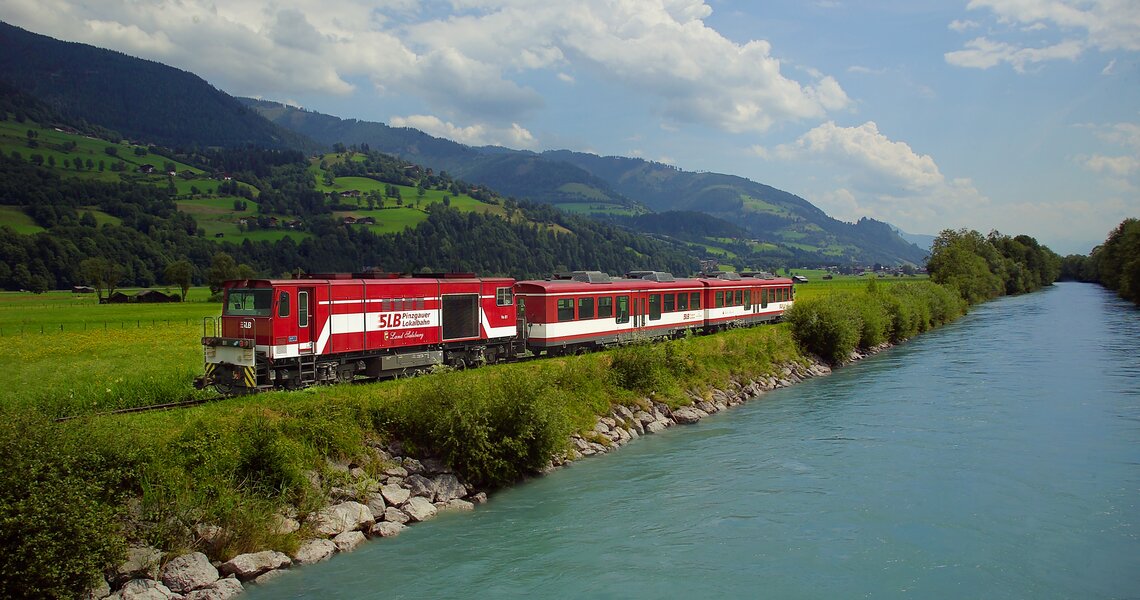 Roter Zug der Pinzgauer Lokalbahn fährt entlang eines Flusses mit grünen Hügeln im Hintergrund. | © Pinzgauer Lokalbahn 