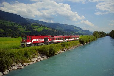 Red train of the Pinzgauer Lokalbahn runs along a river with green hills in the background. | © Pinzgauer Lokalbahn 