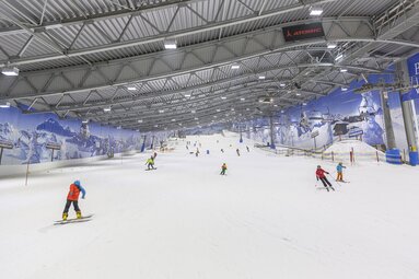 Skihalle mit Menschen, die auf einer Indoor-Piste Ski fahren, umgeben von Alpenmotiven.