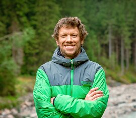 Lächelnder Mitarbeiter von der Nationalparkverwaltung Hohe Tauern in Salzburg, in Nationalparkjacke am Bach. | © NPHT / Glück
