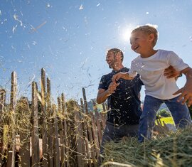 Vater und Kind werfen Heu in die Luft vor einem Holzzaun, sommerlicher Himmel im Hintergrund. | © SalzburgerLand Tourismus