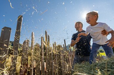 Vater und Kind werfen Heu in die Luft vor einem Holzzaun, sommerlicher Himmel im Hintergrund. | © SalzburgerLand Tourismus