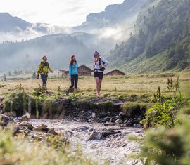 Three hikers walking along a stream in a green valley, surrounded by mist and mountain huts. | © SalzburgerLand Tourismus - Michael Groessinger