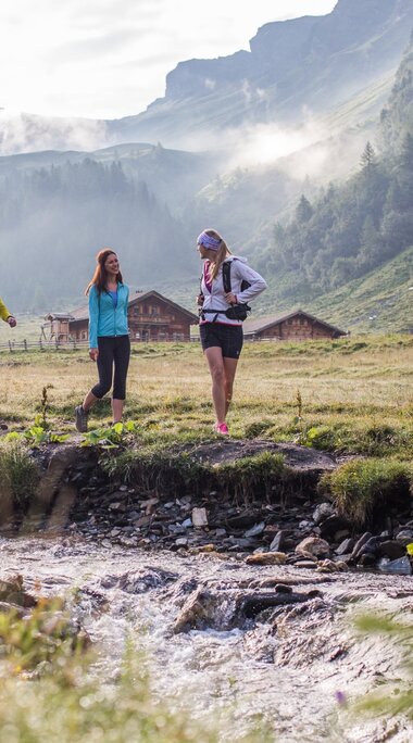 Three hikers walking along a stream in a green valley, surrounded by mist and mountain huts. | © SalzburgerLand Tourismus - Michael Groessinger