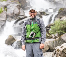 Ein Ranger mit Fernglas und Wanderausrüstung vor einem Wasserfall, strahlend lächelnd. | © SalzburgerLand Tourismus