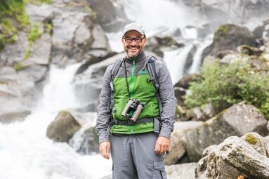 A ranger with binoculars and hiking gear smiling brightly in front of a waterfall. | © SalzburgerLand Tourismus