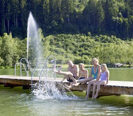 Eltern mit zwei Kindern sitzen am Steg des Badesees und haben Spaß. | © Ferienregion Nationalpark Hohe Tauern - Michael Huber