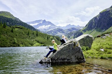Zwei Kinder sitzen auf einem Felsen am Ufer eines Bergsees, umgeben von grünen Hügeln und Bergen. | © Ferienregion Nationalpark Hohe Tauern - Michael Huber