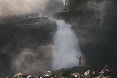 Mann vor einem tosenden Wasserfall, umgeben von aufsteigendem Nebel und Sonnenlichtstrahlen. | © Ferienregion Nationalpark Hohe Tauern - Gerald Demolsky