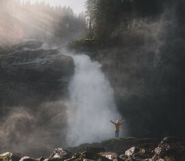 Mann vor einem tosenden Wasserfall, umgeben von aufsteigendem Nebel und Sonnenlichtstrahlen. | © Ferienregion Nationalpark Hohe Tauern - Gerald Demolsky