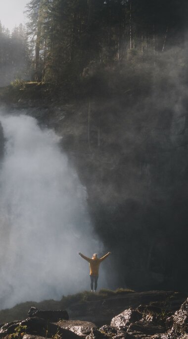 Mann vor einem tosenden Wasserfall, umgeben von aufsteigendem Nebel und Sonnenlichtstrahlen. | © Ferienregion Nationalpark Hohe Tauern - Gerald Demolsky