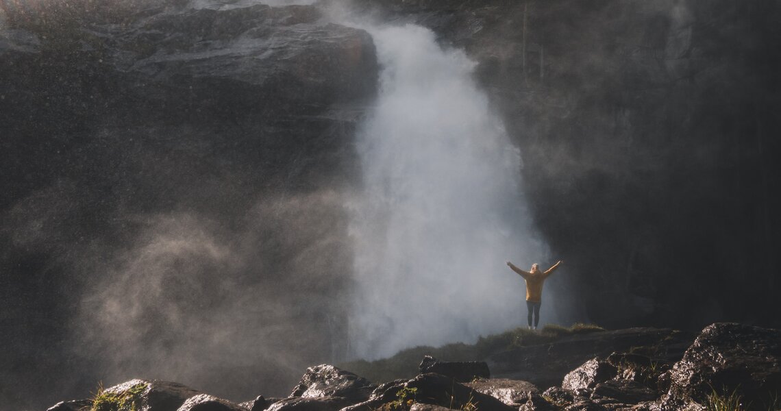 Mann vor einem tosenden Wasserfall, umgeben von aufsteigendem Nebel und Sonnenlichtstrahlen. | © Ferienregion Nationalpark Hohe Tauern - Gerald Demolsky