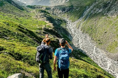 Zwei Wanderer mit Rucksäcken zeigen auf ein hochalpines Tal mit Gletscherblick. | © Ferienregion Nationalpark Hohe Tauern