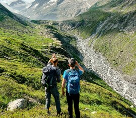 Zwei Wanderer mit Rucksäcken zeigen auf ein hochalpines Tal mit Gletscherblick. | © Ferienregion Nationalpark Hohe Tauern