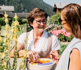 Eine Frau zeigt einer Besucherin Blumen im Kräutergarten, umgeben von alpiner Kulisse. | © Chris Perkles