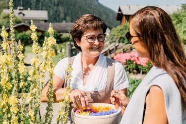 Eine Frau zeigt einer Besucherin Blumen im Kräutergarten, umgeben von alpiner Kulisse. | © Chris Perkles