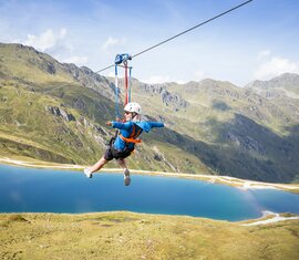 Eine Person gleitet an einer Zipline über einen blauen Bergsee mit beeindruckender Bergkulisse. | © Zillertal Arena